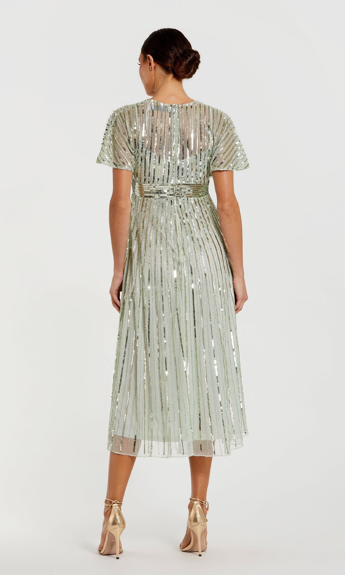 A woman stands facing away in the Mac Duggal 14007, a short sequin wedding-guest dress in light green with vertical metallic stripes and high heels—ideal for semi-formal occasions.